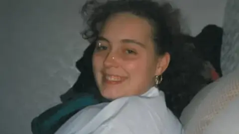 A head and shoulders side-on image of Kellie Pratt, who appears to be sitting on a settee, turning and smiling at the camera. She has brown hair and is wearing a white top and a gold hoop earring. 