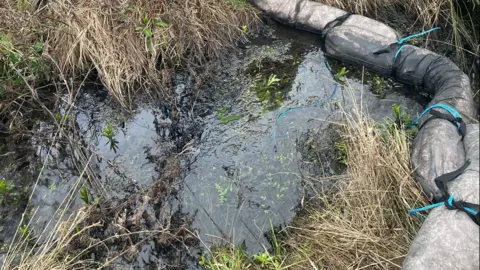 National Highways Oil in a ditch, with a boom to the right. There is grass all around the area.