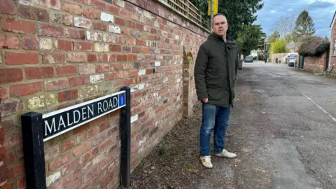 Julian Tooke stands beside a road sign for Malden Road in Cheltenham. He is wearing a khaki coloured coat, blue jeans and white trainers. He looks unimpressed. 