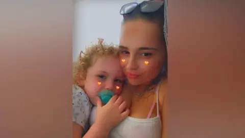 Family handout Destiny and Linnea taking a selfie with a filter over the top. They are both looking at the camera. They have small orange hearts on their cheeks and are cuddling.