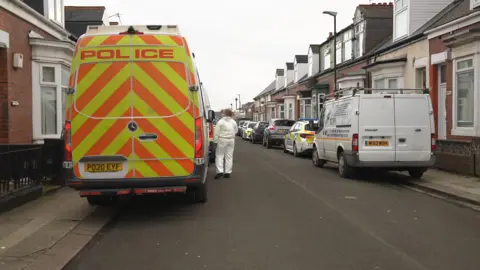 Shot of the back of a police van parked on the left hand side of a road with rows of terraced houses on either side. Next to the van is a person in a white forensic suit. There are parked cars, including a white van, on the other side of the road. 