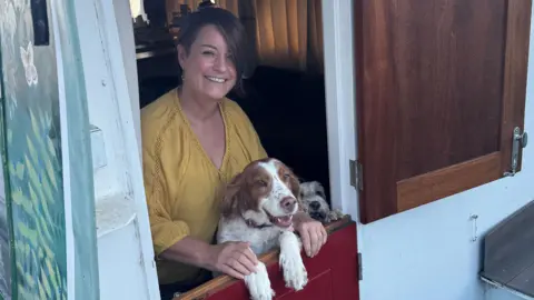 A woman wearing a yellow top standing on and inside her barge with two dogs in front and next to her. She is smiling and looking into the camera.