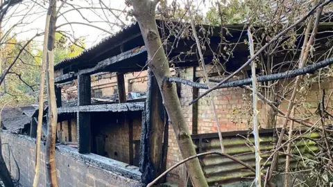 Essex County Fire and Rescue Service A brick building is on the right which has a brick wall leading along the left of the image. Pieces of wood are holding up a tin roof which connects to the building, which has been damaged by the fire.