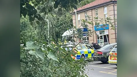 Rachael McMenemy/BBC The front of a shop is damaged with police nearby