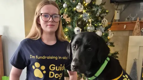 Aniyah, who has long blonde hair and glasses, smiling at the camera, wearing a navy t-shirt with "I'm fundraising in aid of Guide Dogs" written on it in yellow letters. A black Labrador, Spirit, is sat next to her on the right. He has a lime green collar on. They are both sat in front of a decorated Christmas tree.