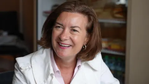 BBC Eluned Morgan wearing a white jacket sat in front of a fridge.