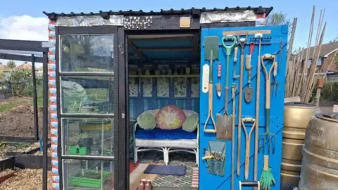 A shed with glass doors, a small sofa chair and tools on the wall. It is in the middle of an allotment.