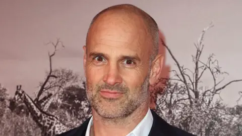 Getty A bald man in a white shirt and pin stripe blazer stands against a wall