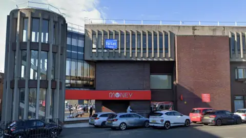 Front elevation of the building showing the circular stairs and branding of Virgin Money visible with couple of cars parked at the front. 