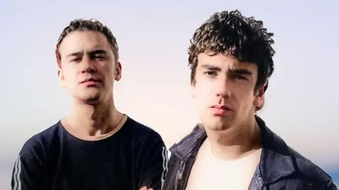 STUART WATRET Carl and Stuart pose for a photo in front of a white wall. They're young, with the picture taken in the 1990s. 