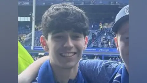 Supplied Sonny has black hair and is wearing an Everton t-shirt and scarf and is smiling at the camera