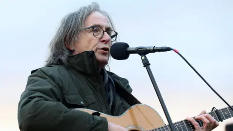 Dougie MacLean is outdoors while singing and playing his guitar. He has shoulder length grey hair, glasses and is wearing a thick green jacket.