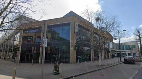 Google A large building with glass windows stretching the length of the facade. A street with bollards and a car parked on it stretches in front of it.
