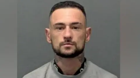 Bedfordshire Police A police custody image of Kieran Humphreys. He has dark hair and a dark beard and is looking straight ahead. He has tattoos on his neck and is wearing a grey top.
