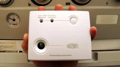 A person holding a white Du Pont carbon monoxide alarm in front of a grey boiler.