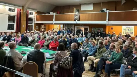 A packed out village hall with rows of people sitting down listening. At the front is a table with people facing the crowd.