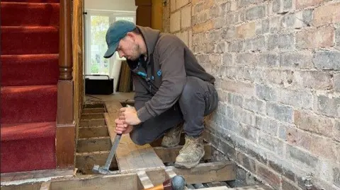 Scott Baggaley Scott Baggaley, a man wearing a black hoodie and black trousers, is using hammer to pull a nail from a floorboard.