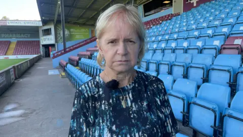 A woman with short blonde hair is pulling a sad expression at a camera. She is wearing a dark blue to with lighter blue on it and is stood in a football stadium next to several rows of blue and red chairs.