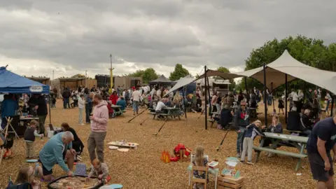 Seb A large group of people at a food event, with benches and tents. There is gravel on the ground. 