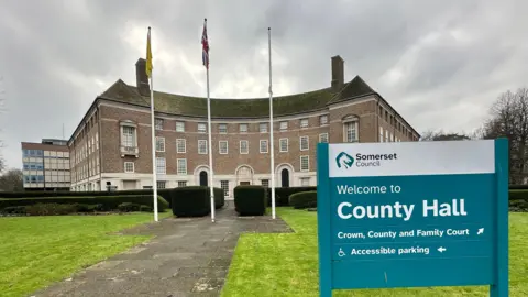 Somerset Council's county hall building from the front. It is brown, curved and four-storeys high. There is a sign in the foreground saying Welcome to County Hall. Three flag poles are in front of the building.