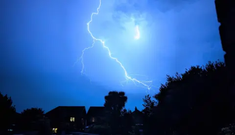 Hengist of Woky Two bolts of lightning shoot across a bright blue sky above houses and trees on the horizon.