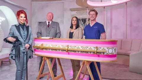 Channel 4 Hayley McCaughran, who has dyed red hair tied back and is wearing a light blue suit, stands next to a pink striped coffin holding a black cane. Next to her are her colleague, a bald man in a light grey suit, and a woman with black hair and a man in blue scrubs. 