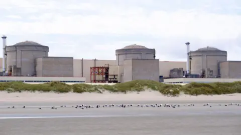 Getty Images A still image of the Gravelines nuclear power plant, with three stone-coloured units connected by a large white wall, set behind a stretch of empty coast with the tide out.