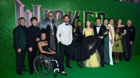 Getty Images (L-R) Ethan Slater, Marissa Bode, Jeff Goldblum, Jonathan Bailey, Cynthia Erivo, Ariana Grande, Michelle Yeoh, Bowen Yang, Karis Musongole and Bronwyn James, all in smart evening wear, stand on a green carpet and in front of a green background carrying the word Wicked