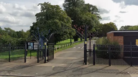 Google A park with bright green grass, and tall full leafy trees. There are black railings at the front of the park with a blue and red swirly pattern creating an archways on the entrance 