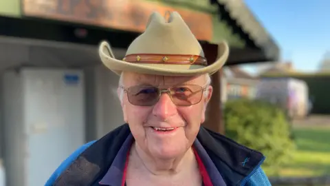 Gerry Smith smiles at the camera. He wears a light green cowboy hair, blue coat and purple polo t-shirt underneath.