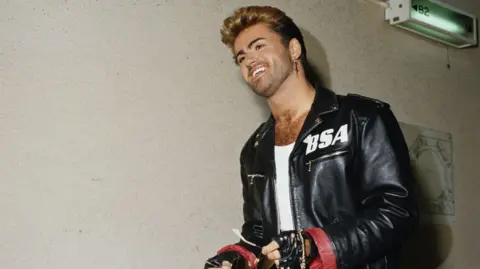 George Michael wearing a black jacket with a white BSA logo on the left side.
