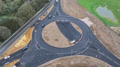 An aerial image of a roundabout under construction. It is surrounded by trees, greenery, and mud.