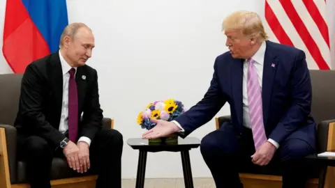 Reuters Russian leader Vladimir Putin, on the left, looks down at the hands of Donald Trump, who is speaking to him on the right. Both men are sitting in suits and holding flags behind them. There are some flowers on the table between them.