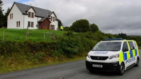 PA Media A police van on the road outside the home of the MacKinnons, where John MacKinnon was shot dead by his brother in law. The home has white walls, with a brown doorway and entrance, while there is greenery in a large garden outside it. 