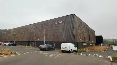 The Graham Sports Centre at Maiden Castle, Durham University Sports Complex. It is a large industrial unit that has brown panelled cladding. there are several cars and vans parked outside. It is a grey cloudy day.