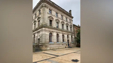 A sandstone grand Victorian era building in Derry's Guildhall Square stands over four floors. Steps leading up to the building are seen on the left.
