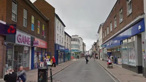 Google image of Barnstaple High Street. Retailers including Poundland and Boots can be seen along the street. People are walking along the pavement.