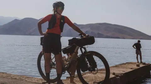 A women is standing with her bike at the edge of a large body of water and behind her you can see mountains. She is wearing cycling gear and a helmet and looking into the distance smiling. 