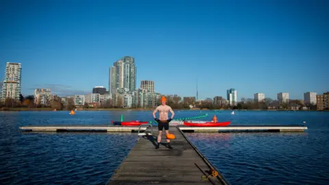 Emma Lynch/BBC James stands on a ramp out across the west reservoir. He is wearing an orange bobble hat and swimming shorts as well as swimming socks. The sky is blue and there are high rise buildings in the distance, beyond the water's edge.