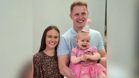 Rebecca and Harry Foster with their baby daughter, Olivia. Rebecca has long dark hair and is looking directly at the camera and smiling. She is standing next to Harry. He has blonde short hair and is wearing a light blue polo shirt. He is holding Olivia. She has blonde short hair and is wearing a pink and white stripe top with a pointed party hat.