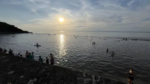 Clevedon Marine Lake pictured at sunset. The marine lake is full and people are swimming, standing and also riding paddleboards