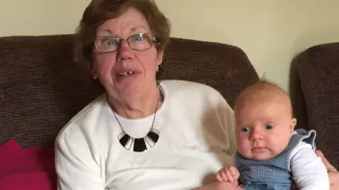 Family photo Kathleen McGarvey, a pensioner with short brown hair, is sitting on a brown sofa holding a baby.  She is wearing glasses, a white top and a black and white necklace.  The baby has short, blonde hair and is wearing a blue and white outfit.