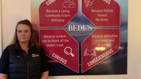 Dani Maynard Dani has long brown hair. She stands in front of the logo of the School of St. Bede showing the words compassionate, courageous, curious and conscientious