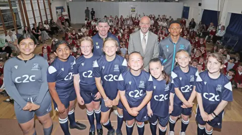 c2c The girls, still wearing their blue kit, stand with their head teacher, Kevin Maher and Nathan Ralph on a stage inside their school hall. They are smiling and are in front of all their fellow school pupils, who are sitting on the floor.