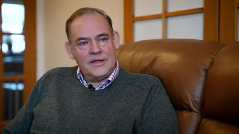 A man with short brown hair, wearing a checked shirt and dark grey jumper, sitting on a brown settee in his living room