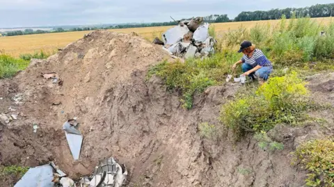 Royal Agricultural University A woman sits on the edge of a large bomb crater in a field in Ukraine. In the background a large field of a light-coloured crop is visible