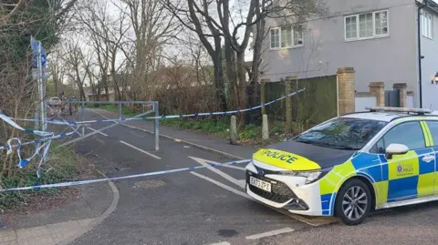 A police car partially blocks the entrance to a cycle path, which has also been cordoned off with blue and white tape. Trees line both sides of the path.