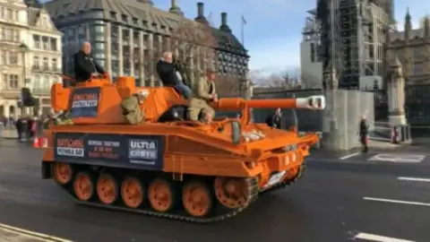 An orange tank with three men sitting on top of it in Parliament Square, London. The tank has a gun mounted on the front.