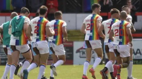Keighley Cougars A group of Keighley players run away from the camera on the pitch. They're wearing white jerseys adorned with Pride flags.