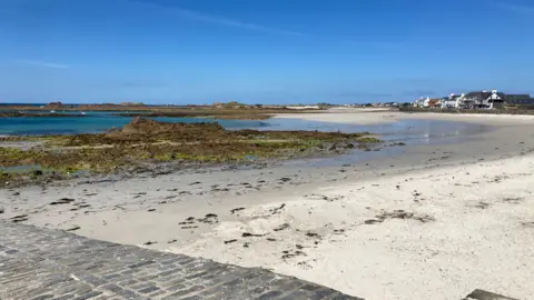 The photo shows a wide sandy beach at low tide, with shallow pools of water, exposed rocky outcrops and patches of green seaweed stretching across the shoreline. In the foreground there is a stone-paved promenade, while in the distance a row of coastal houses lines the edge of the bay beneath a bright blue sky with only a few wispy clouds visible.